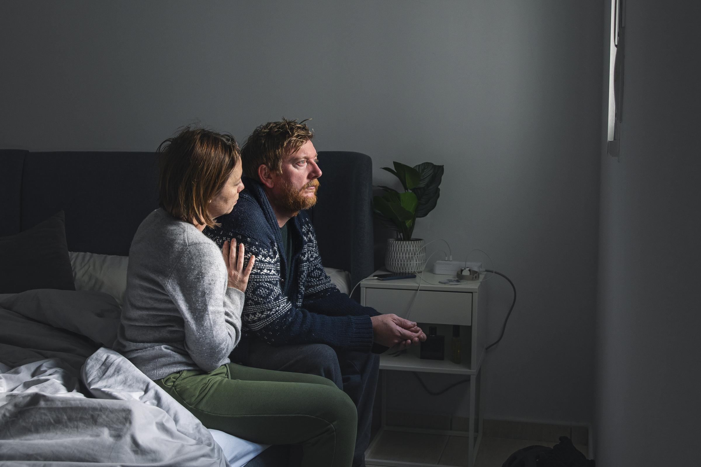 Powerful portrait of a mature couple sitting on their bed. The man looks sad. The woman is comforting him.