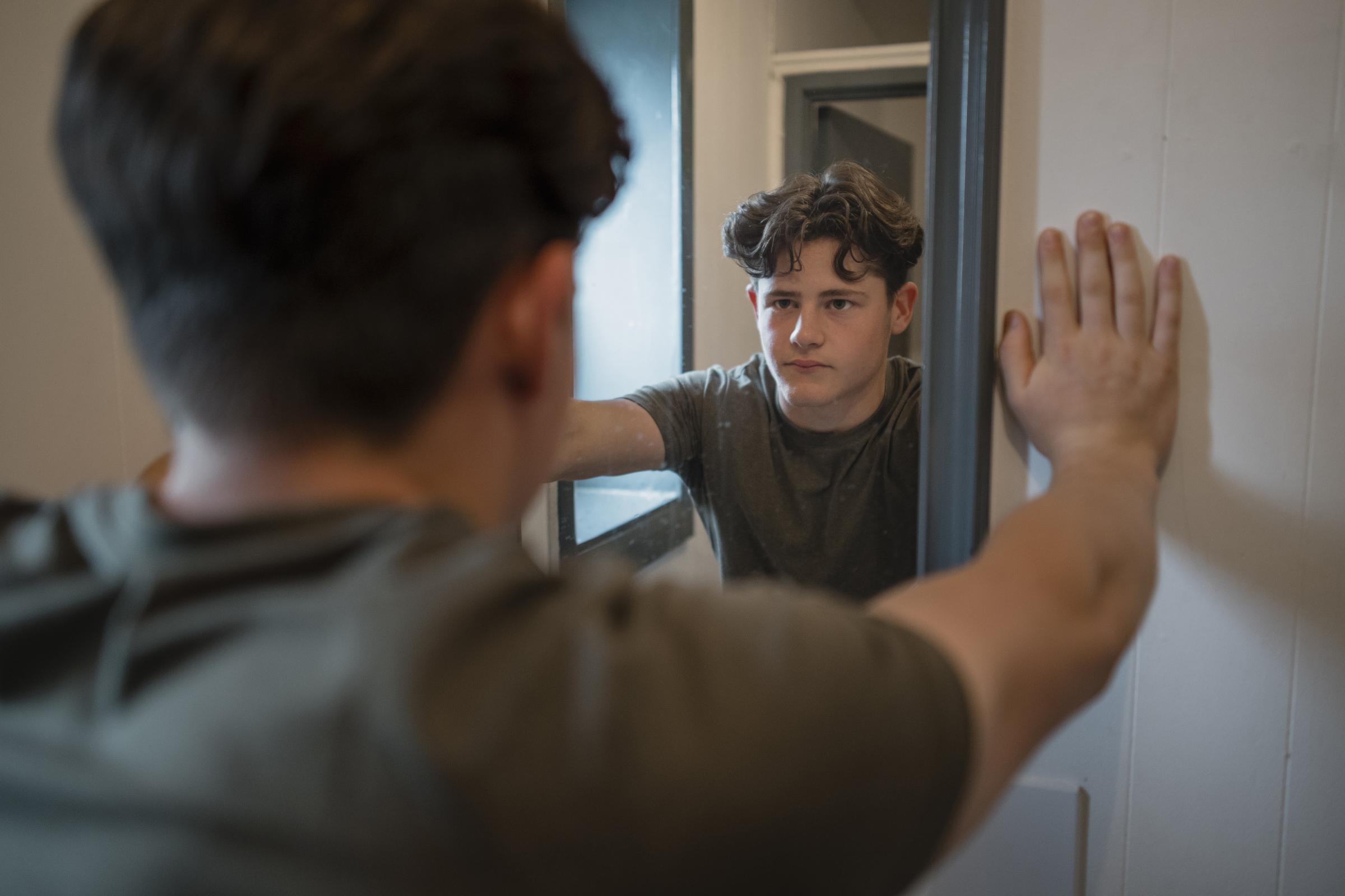 An over-the-shoulder shot of a young teenage male athlete in the gym changing rooms, he is taking a break from his gym session to catch his breath, he has his hands on the wall and is looking at his reflection in the mirror..