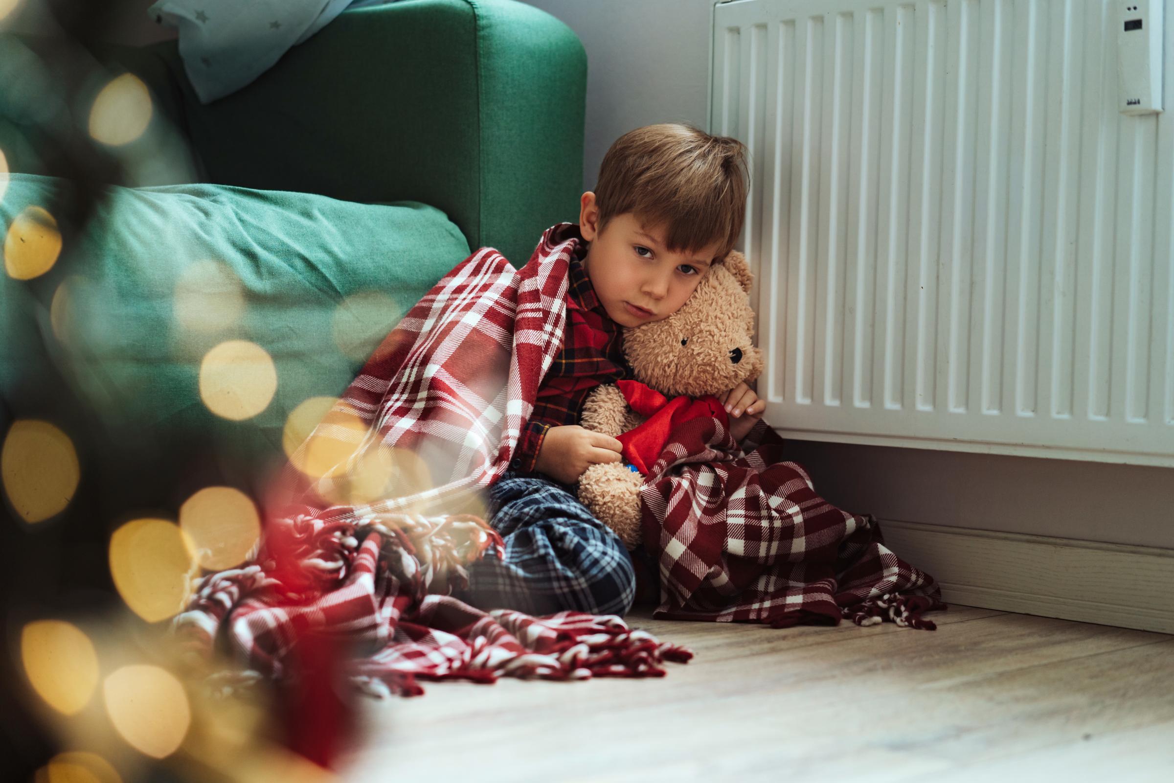 cute little boy wrapped id plaid falling asleep by heater hugging teddy bear in santa hat. Christmas time. Cold at home, Energy crisis. Fuel poverty